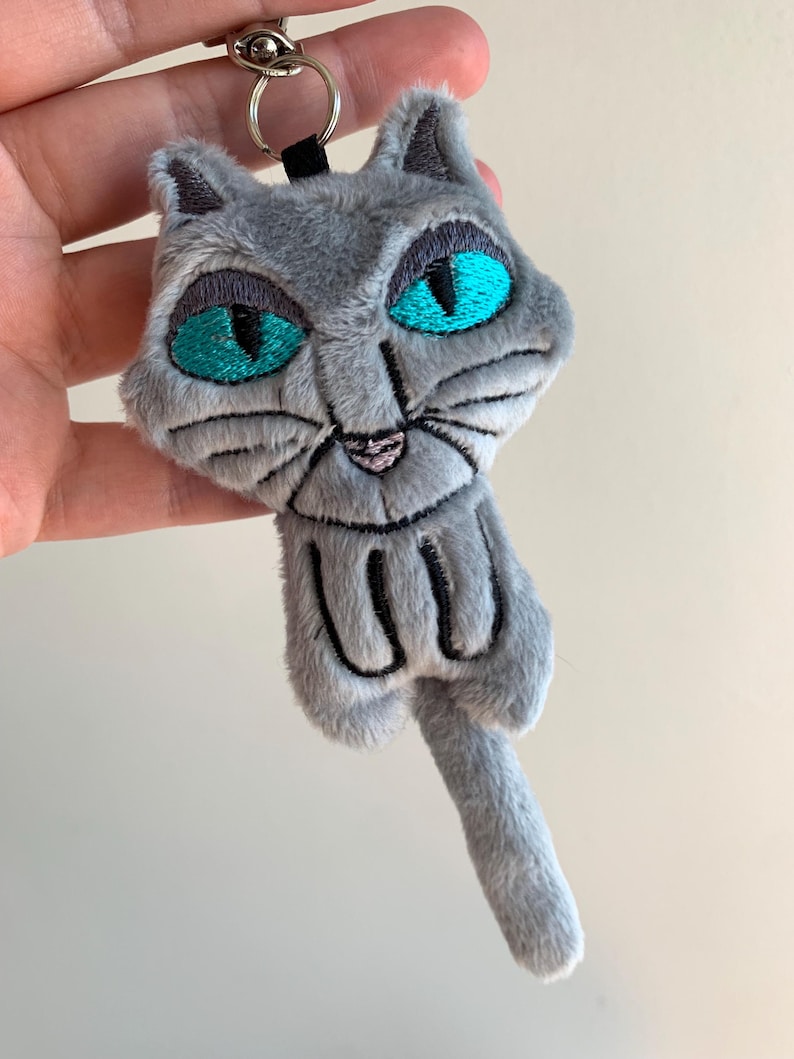 May include: A gray plush cat keychain with embroidered details. The cat has large teal eyes, a pink nose, and black whiskers. The keychain includes a silver metal ring and a black fabric loop.