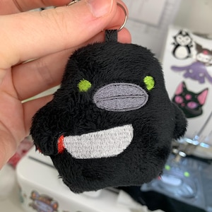 Peut inclure: Un porte-clés en peluche noire en forme d'oiseau arrondi. La peluche présente des yeux brodés vert citron, un bec ovale gris et un couteau blanc avec un manche rouge tenu dans son aile. Un anneau en métal argenté est fixé au sommet.