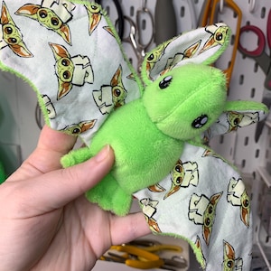 May include: A plush green creature with large black eyes and patterned wings. The wings are white with a repeating image of a cartoon character. The edges of the wings are trimmed with green fabric. The plush toy is held in a person's hand.