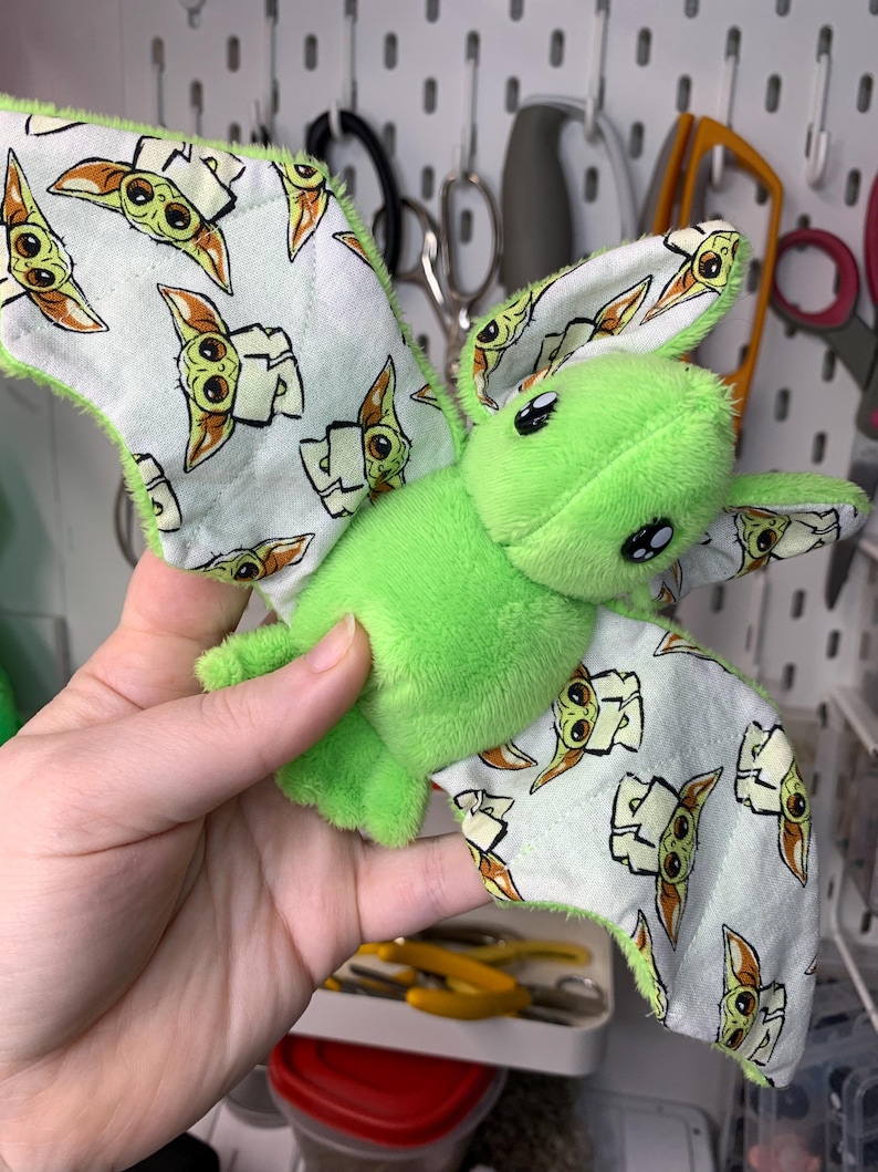 May include: A plush green creature with large black eyes and patterned wings. The wings are white with a repeating image of a cartoon character. The edges of the wings are trimmed with green fabric. The plush toy is held in a person's hand.