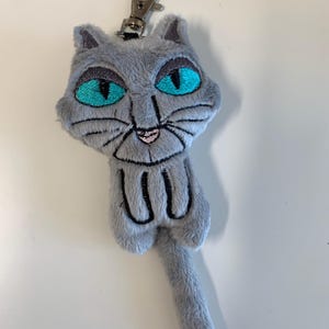 May include: A gray plush cat keychain with bright turquoise eyes and black embroidered details. The cat has a silver metal clasp for attaching to keys or a bag. The cat's tail is long and slender.