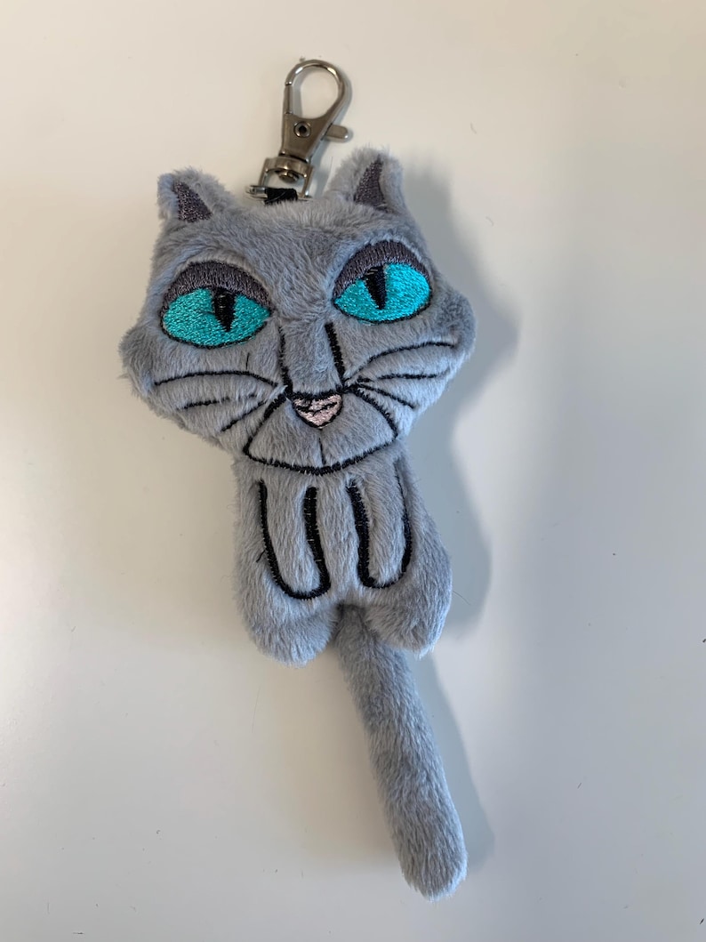 May include: A gray plush cat keychain with bright turquoise eyes and black embroidered details. The cat has a silver metal clasp for attaching to keys or a bag. The cat's tail is long and slender.