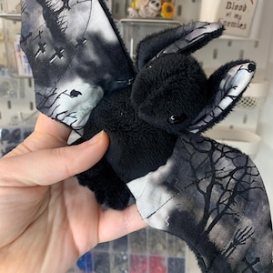 Peut inclure: Une chauve-souris en peluche noire avec des ailes ornées d'une scène de cimetière avec des croix et des arbres. Le corps est doux et duveteux, parfait pour Halloween ou une décoration gothique.