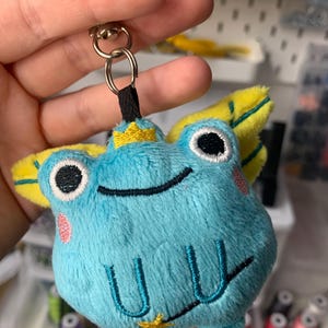 May include: A light blue plush frog keyring with yellow wings and a gold crown. The frog features black eyes, pink cheeks, and a stitched smile. The keyring includes a silver clasp and a black fabric loop.