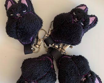 May include: Four black cat-shaped keychains with purple embroidered details and pink inner ears. Each plush cat has a silver-colored clasp and a black fabric loop for attachment. The keychains are arranged in a circular pattern.