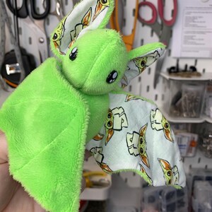 May include: A plush green bat with large black eyes and patterned wings. The wings feature a light blue fabric with a repeating cartoon character design. The bat's body is a soft, fuzzy material.
