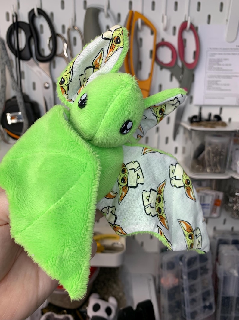 May include: A plush green bat with large black eyes and patterned wings. The wings feature a light blue fabric with a repeating cartoon character design. The bat's body is a soft, fuzzy material.