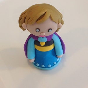 May include: A small, hand-crafted figurine of a girl with blonde hair, wearing a blue dress with purple and yellow accents. The figurine is made of fondant and is likely a cake topper.