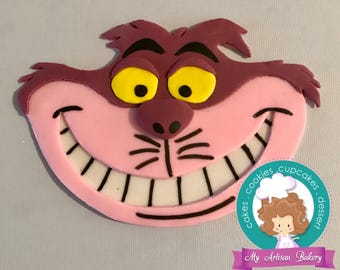 May include: A pink and purple Cheshire Cat face with a wide grin and yellow eyes. The cat's mouth is open, revealing white teeth with black outlines. The face is made of fondant and is likely a cake topper.