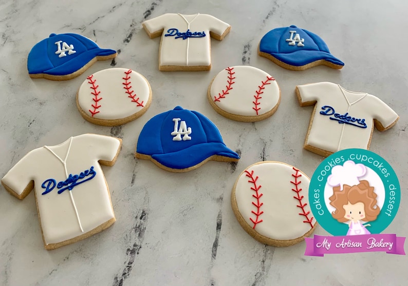 Pu&ograve; includere: Un set di nove biscotti di zucchero a tema baseball decorati con glassa blu e bianca. I biscotti raffigurano palle da baseball, maglie da baseball e cappellini da baseball con il logo dei LA Dodgers.