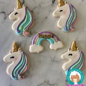 May include: Five decorated sugar cookies, four are unicorn-shaped with pastel blue, green, and purple icing, and one is a rainbow with clouds and the name "Maggie" in gold icing. The cookies are on a white marble surface.