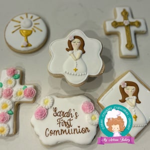 May include: Six decorated sugar cookies for a First Communion celebration. The cookies feature a chalice, a cross with pink flowers, a cross with gold accents, and three cookies with a girl in a white dress praying. One cookie has the text "Sarah's First Communion" on it.