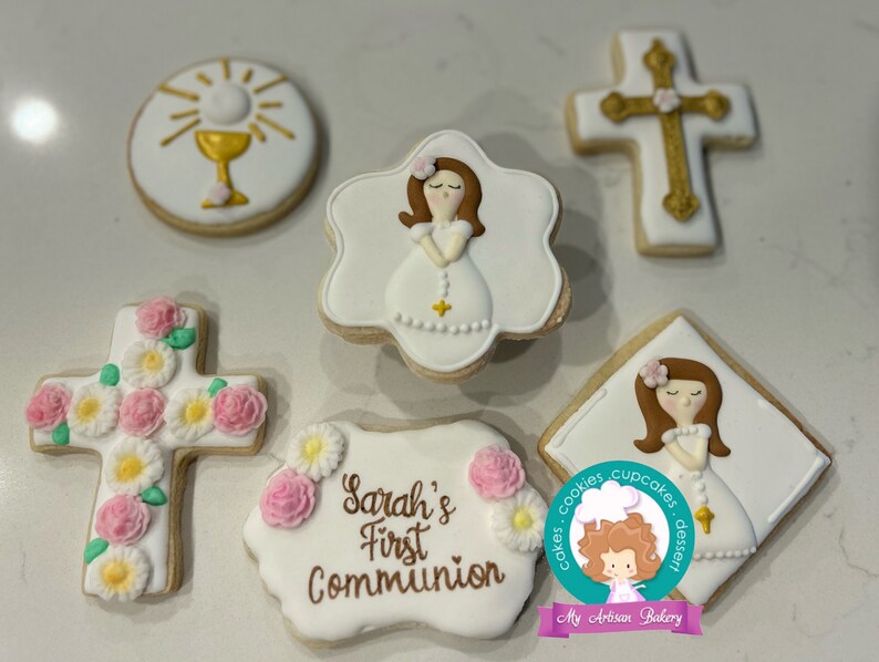 May include: Six decorated sugar cookies for a First Communion celebration. The cookies feature a chalice, a cross with pink flowers, a cross with gold accents, and three cookies with a girl in a white dress praying. One cookie has the text "Sarah's First Communion" on it.