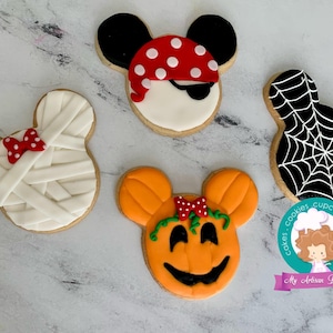 May include: Four decorated sugar cookies shaped like Mickey Mouse. The cookies are decorated with different Halloween themes: a mummy, a pirate, a pumpkin, and a spiderweb. The cookies are on a white marble surface. The text "cakes - cookies - cupcakes - desserts" is on a teal circle with a pink border. The text "My Artisan Bakery" is below the circle.