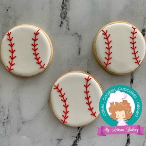 Pu&ograve; includere: Tre biscotti al burro a tema baseball, bianchi e rossi, con dettagli di cuciture rosse. I biscotti sono su uno sfondo grigio.