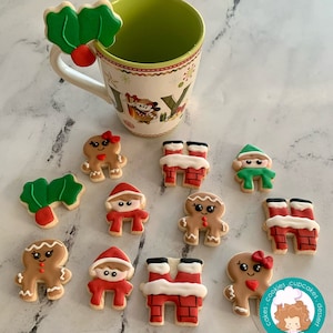 May include: A collection of Christmas-themed sugar cookies decorated with icing. The cookies feature gingerbread people, Santa Claus, elves, and chimneys. The cookies are arranged on a white surface with a green mug in the background.