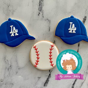 Pu&ograve; includere: Tre biscotti di zucchero decorati: due cappellini da baseball blu con logo "LA" bianco e una palla da baseball bianca con cuciture rosse.