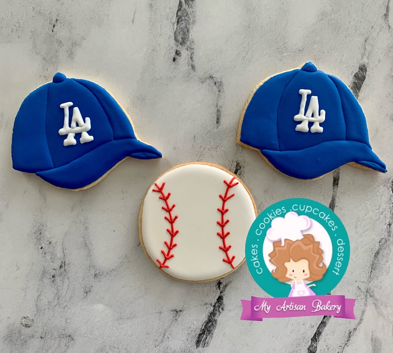 Pu&ograve; includere: Tre biscotti di zucchero decorati: due cappellini da baseball blu con logo "LA" bianco e una palla da baseball bianca con cuciture rosse.
