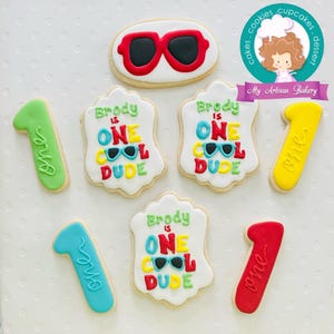 May include: A collection of decorated sugar cookies. The cookies include a pair of sunglasses, the number one, and cookies that say "Brody is ONE COOL DUDE" in colorful letters. The cookies are on a white surface.
