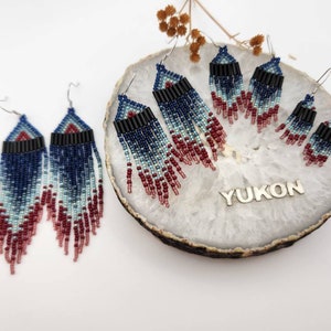 Peut inclure: Un ensemble de quatre paires de boucles d'oreilles en perles avec un dégradé de couleurs bleu, turquoise et bordeaux. Les boucles d'oreilles sont faites de petites perles de rocaille et ont un design en frange. Les boucles d'oreilles sont présentées sur un fond blanc avec une dalle de pierre et le mot "YUKON" écrit sur la dalle.