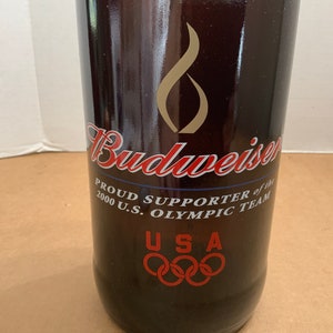 May include: A brown glass bottle with a Budweiser label. The label says "Proud Supporter of the 2000 U.S. Olympic Team" and features the Olympic rings and the letters "USA".