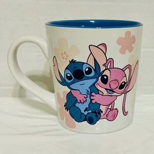 May include: White ceramic mug with a blue interior and a white handle. The mug features a cartoon illustration of a blue and pink alien-like characters hugging, with pink flower accents.