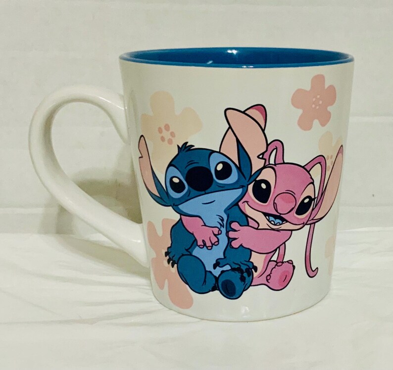 May include: White ceramic mug with a blue interior and a white handle. The mug features a cartoon illustration of a blue and pink alien-like characters hugging, with pink flower accents.