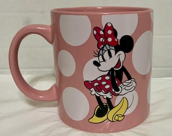 May include: Pink ceramic mug with white polka dots and a Minnie Mouse graphic. The mug features a pink handle and a smiling Minnie Mouse wearing a red polka dot bow and dress. Yellow shoes complete the look.