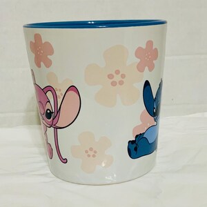 May include: White ceramic mug with a blue rim, featuring pink floral designs and cartoon characters Stitch and Angel. The mug is cylindrical and has a smooth surface.