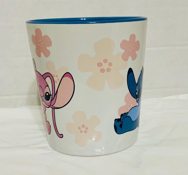 May include: White ceramic mug with a blue rim, featuring pink floral designs and cartoon characters Stitch and Angel. The mug is cylindrical and has a smooth surface.