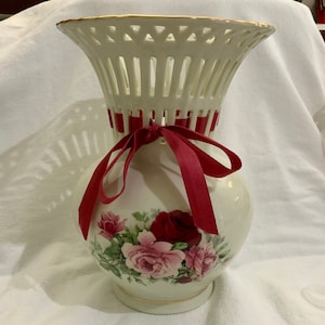 May include: White ceramic vase with a red ribbon and floral design. The vase has a lattice-like top and a round body with a floral pattern featuring pink and red roses.