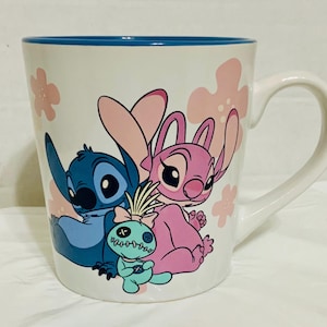 May include: White ceramic mug featuring Stitch, Angel, and a small doll. The mug has a blue rim and a white handle. Pink flower designs are scattered around the characters. The characters are in shades of blue and pink.
