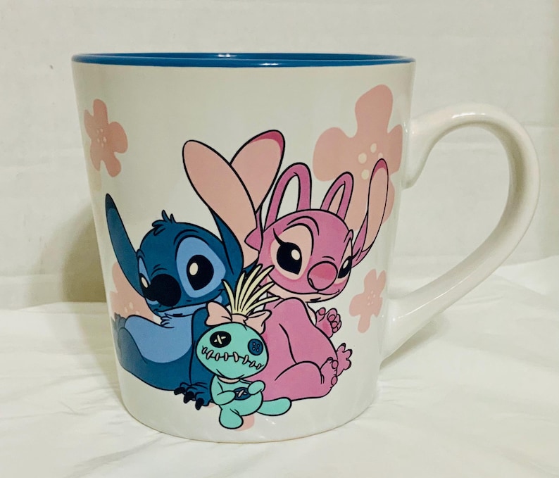 May include: White ceramic mug featuring Stitch, Angel, and a small doll. The mug has a blue rim and a white handle. Pink flower designs are scattered around the characters. The characters are in shades of blue and pink.