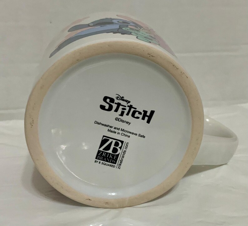 May include: White ceramic mug featuring the Disney character Stitch. The mug has a handle and a design of Stitch on the side. The bottom of the mug has the Disney Stitch logo and text indicating it is dishwasher and microwave safe.