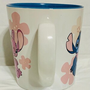 May include: White ceramic mug with a blue interior and a white handle. The mug features pink and white floral designs and illustrations of the characters Stitch and Angel from the animated film Lilo & Stitch.