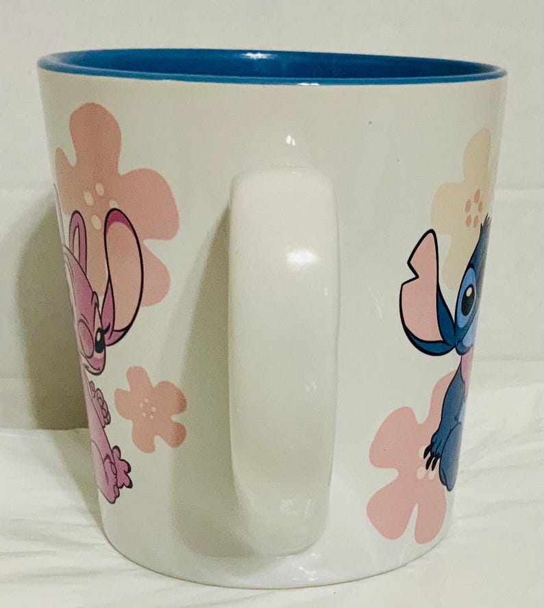 May include: White ceramic mug with a blue interior and a white handle. The mug features pink and white floral designs and illustrations of the characters Stitch and Angel from the animated film Lilo & Stitch.