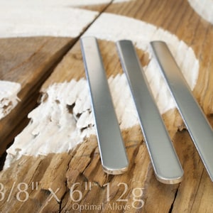 May include: Three silver metal bars, each measuring 3/8 inch by 6 inches, are arranged on a wooden surface. The bars are smooth and have rounded edges. The text "3/8" x 6" 12g" is visible on the wooden surface.