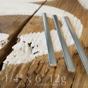 May include: Three silver metal rods, each 1/4 inch thick and 6 inches long, on a wooden surface. The text "1/4" x 6" 12g" is printed below the rods. The text "Optimal Alloys" is printed below the measurement text.