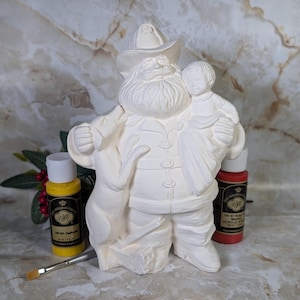 May include: An unpainted ceramic figurine of a bearded figure wearing a hat and coat, holding a small child in one arm and a dog at their side. The figurine is white and matte, accompanied by a small paintbrush and two bottles of acrylic paint.