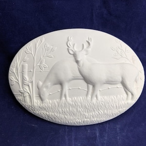 May include: Oval-shaped, white ceramic plaque featuring a detailed relief of two deer in a grassy field. One deer grazes, while the other stands with antlers. A fence and foliage are also depicted.