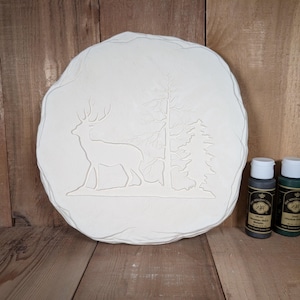 May include: A round, off-white stone-like plaque with a raised relief of a deer and trees. Two small paint bottles, one dark gray and one dark green, are next to the plaque. The plaque is approximately 12 inches in diameter.