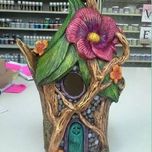 May include: A whimsical fairy house made of sculpted wood and painted with vibrant colors. The house features a large pink flower, a green door, and a birdhouse opening. The house is decorated with small stones and leaves.