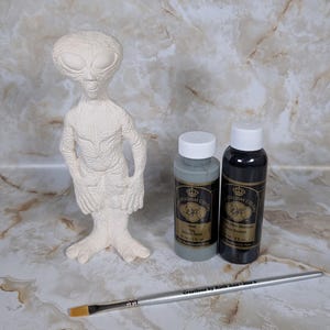 May include: A white alien figurine with a textured surface, standing next to two bottles of paint and a paintbrush. The bottles are labeled with the brand "Holiday Colors". The background is a marble-like surface.