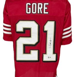 May include: Red football jersey with the name "GORE" and the number "21" in white with black and gold trim. The jersey has white stripes on the sleeves and a signature on the number.