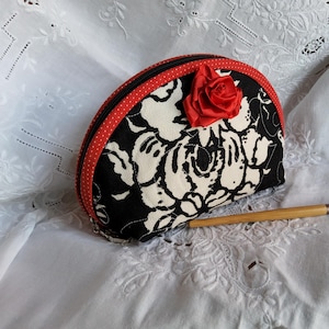 May include: A black and white floral print cosmetic bag with a red trim and a red rose accent. A makeup brush with a wooden handle is next to the bag. The bag is on a white, embroidered surface.