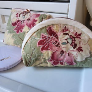 May include: Two floral print cosmetic bags and a compact. The bags have a cream-colored zipper and a quilted floral pattern in shades of pink, green, and cream. The compact is white with the word "fancil" printed on it.