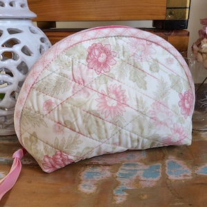 May include: A quilted, half-moon shaped tea cozy with a floral pattern in pink and green on a white background. The cozy has a pink ribbon loop and is placed on a distressed wooden surface.