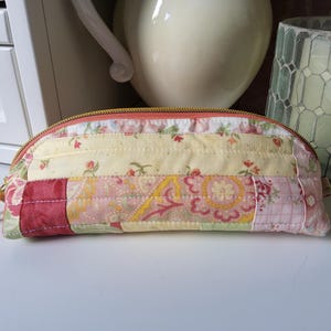 May include: A semi-circular quilted pencil case featuring a patchwork design of floral and paisley patterns in shades of yellow, pink, and red. The pouch is secured with a gold-toned metal zip along the top edge and has a soft, padded texture.