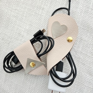 May include: Two beige leather cord organizers with gold accents. One organizer has a heart-shaped patch and the other has a triangular patch. Both organizers are holding black cords.