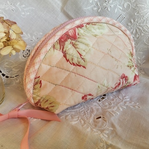 May include: A quilted, half-moon shaped cosmetic bag with a floral pattern in red, green, and cream. The bag has a pink zipper and a matching pink ribbon attached. The bag is on a white lace surface.